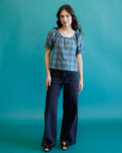 A young woman modeling a blue and gray plaid puff-sleeve top with dark wide-leg pants, standing against a solid teal studio backdrop.