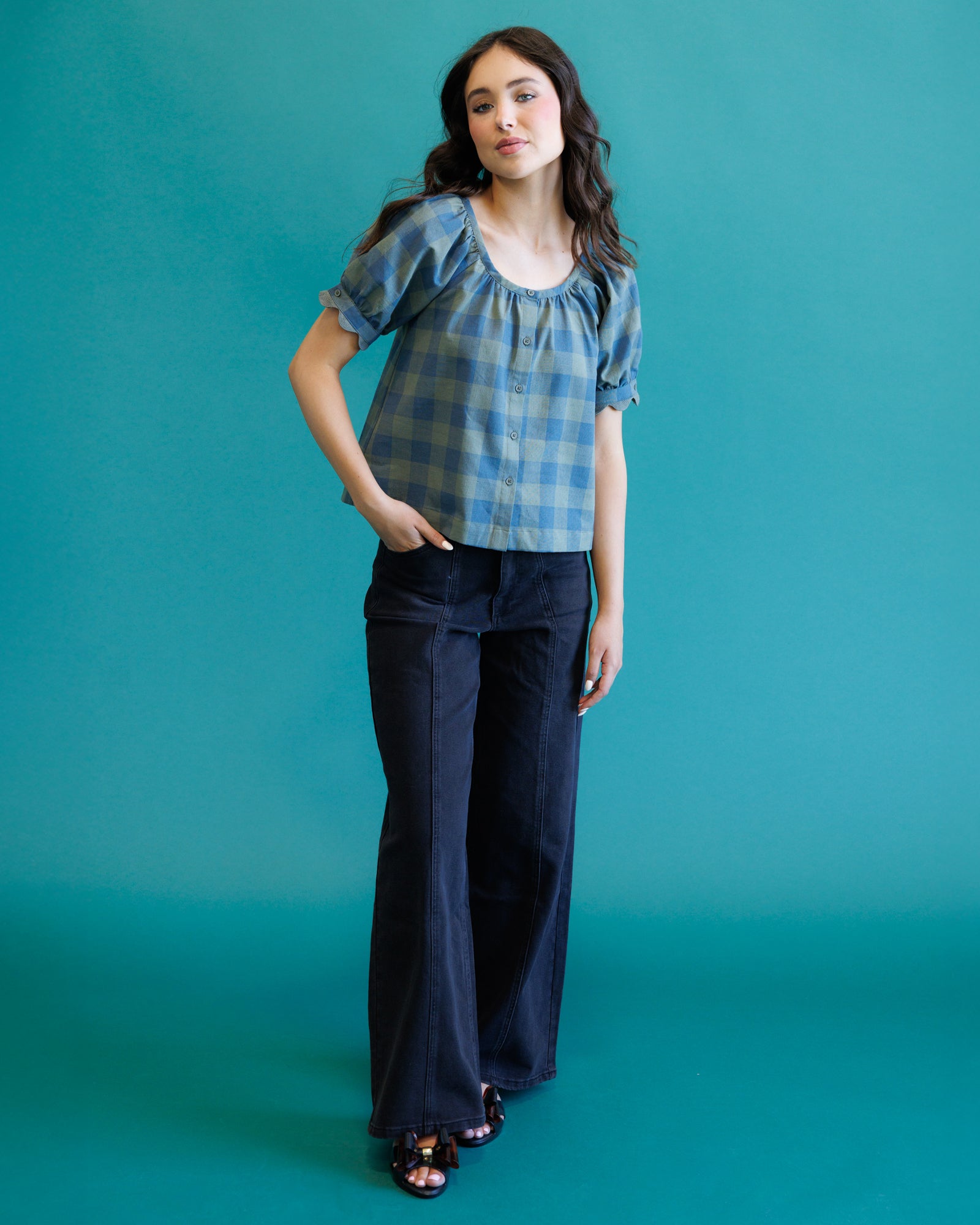 A woman modeling a blue and gray plaid puff-sleeve top with dark wide-leg jeans, standing against a solid teal studio backdrop.