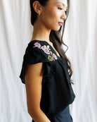 Woman wearing a black top with floral embroidery against a white curtain background