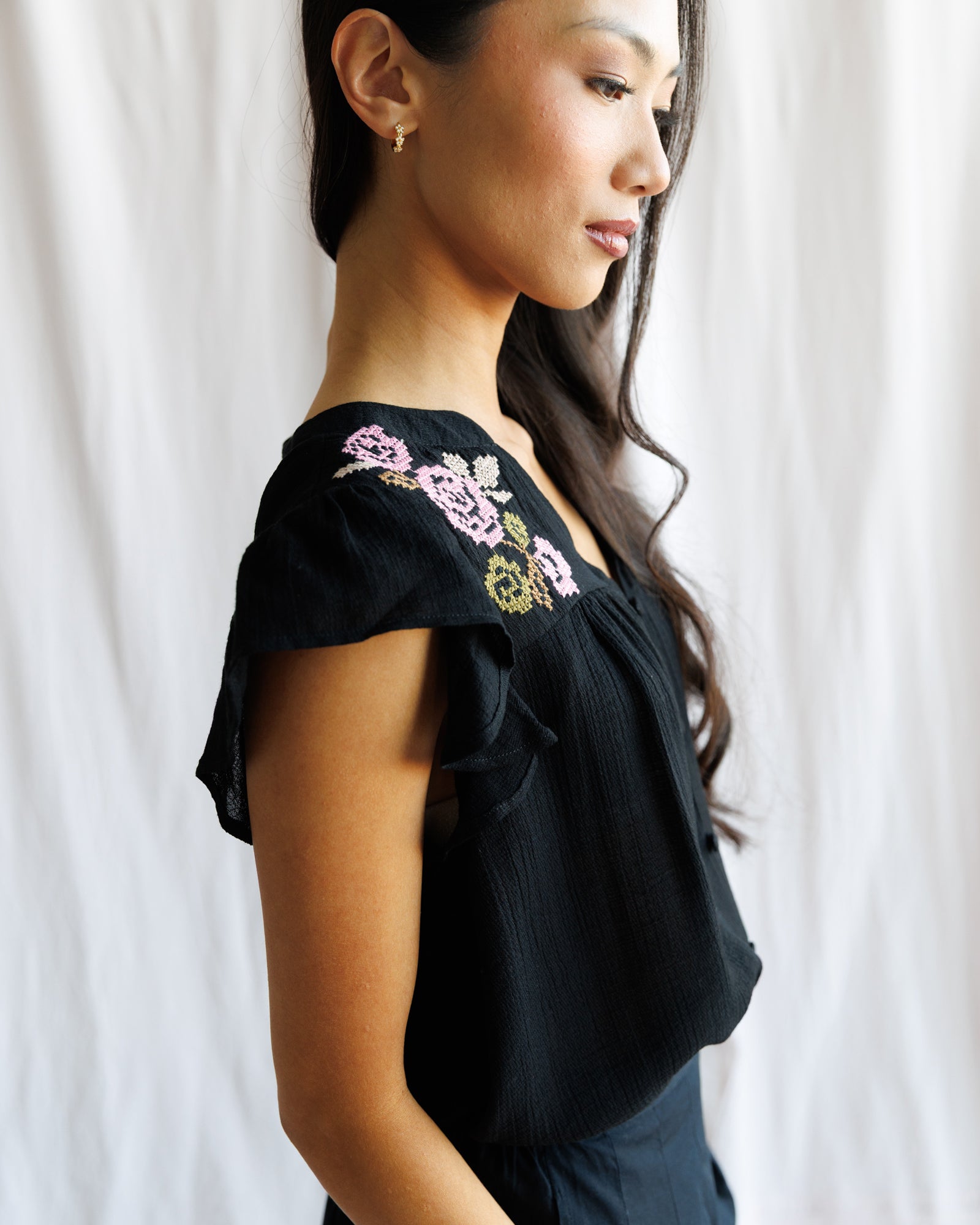 Woman wearing a black top with floral embroidery against a white curtain background