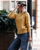 A woman wearing a mustard yellow cable-knit sweater, blue jeans, and a black baseball cap with white lettering, standing outdoors next to a brick wall and greenery.