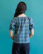 A woman viewed from behind wearing a blue and light gray plaid puff-sleeve blouse and dark jeans, standing against a solid teal background.