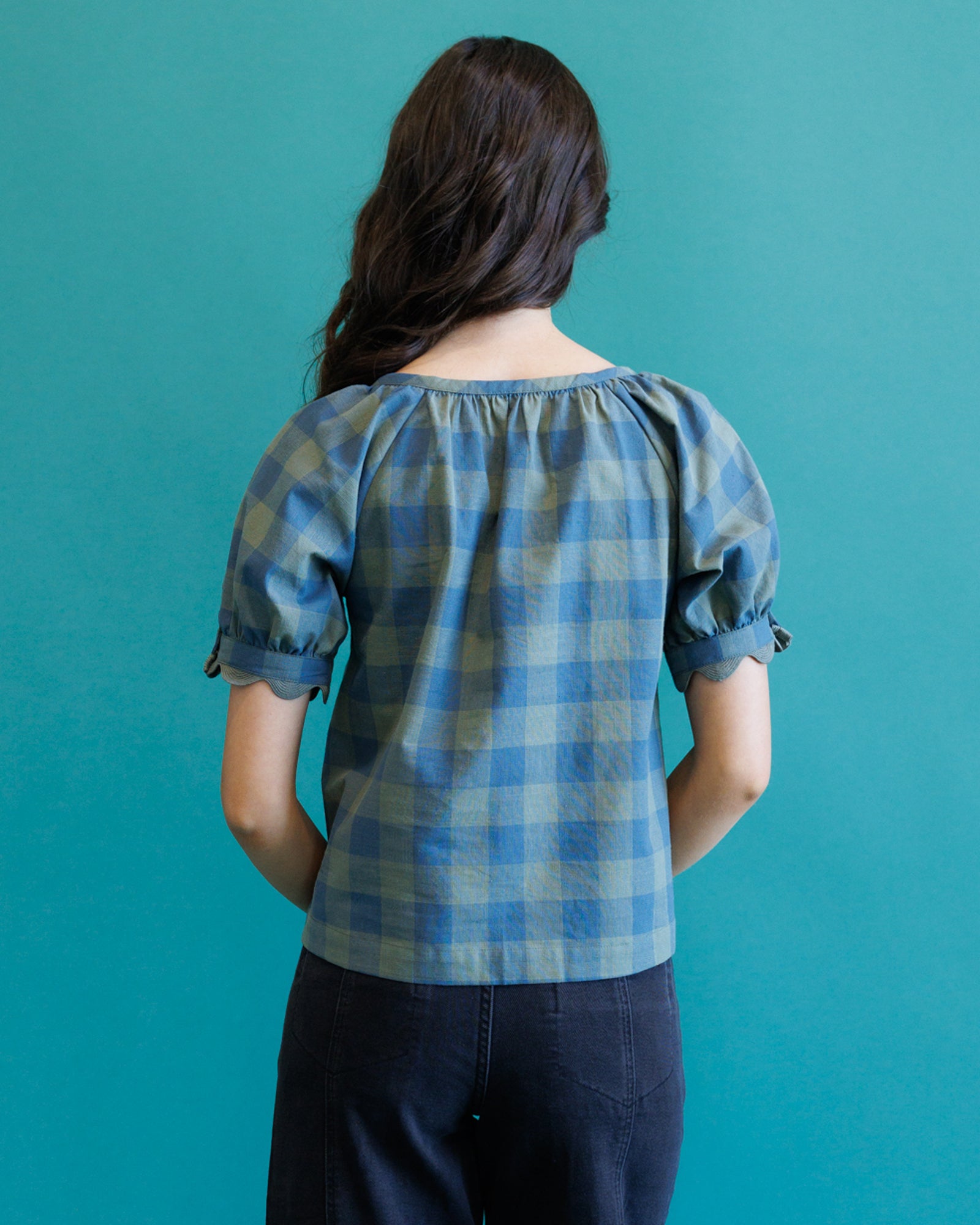 A woman viewed from behind wearing a blue and light gray plaid puff-sleeve blouse and dark jeans, standing against a solid teal background.