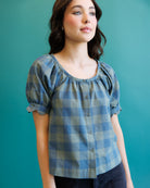 A woman wearing a blue and gray plaid puff-sleeve blouse with a button-down front, posed against a solid teal background.