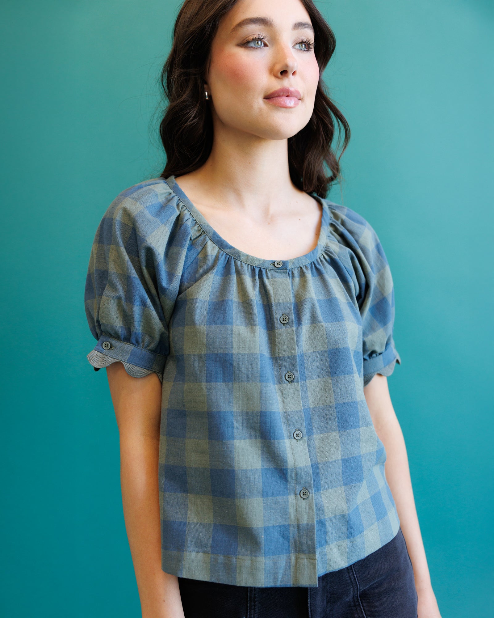 A woman wearing a blue and gray plaid puff-sleeve blouse with a button-down front, posed against a solid teal background.