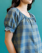 A close-up of a person wearing a blue and gray gingham blouse with puff sleeves and a button front, set against a solid teal background.