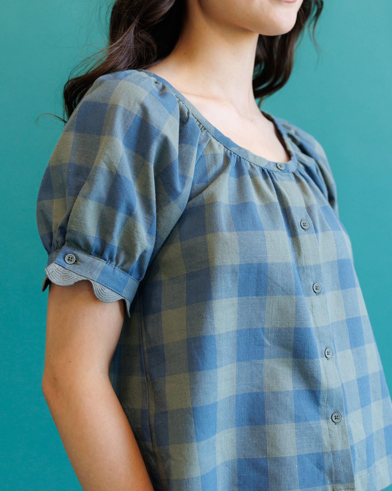 A close-up of a person wearing a blue and gray gingham blouse with puff sleeves and a button front, set against a solid teal background.