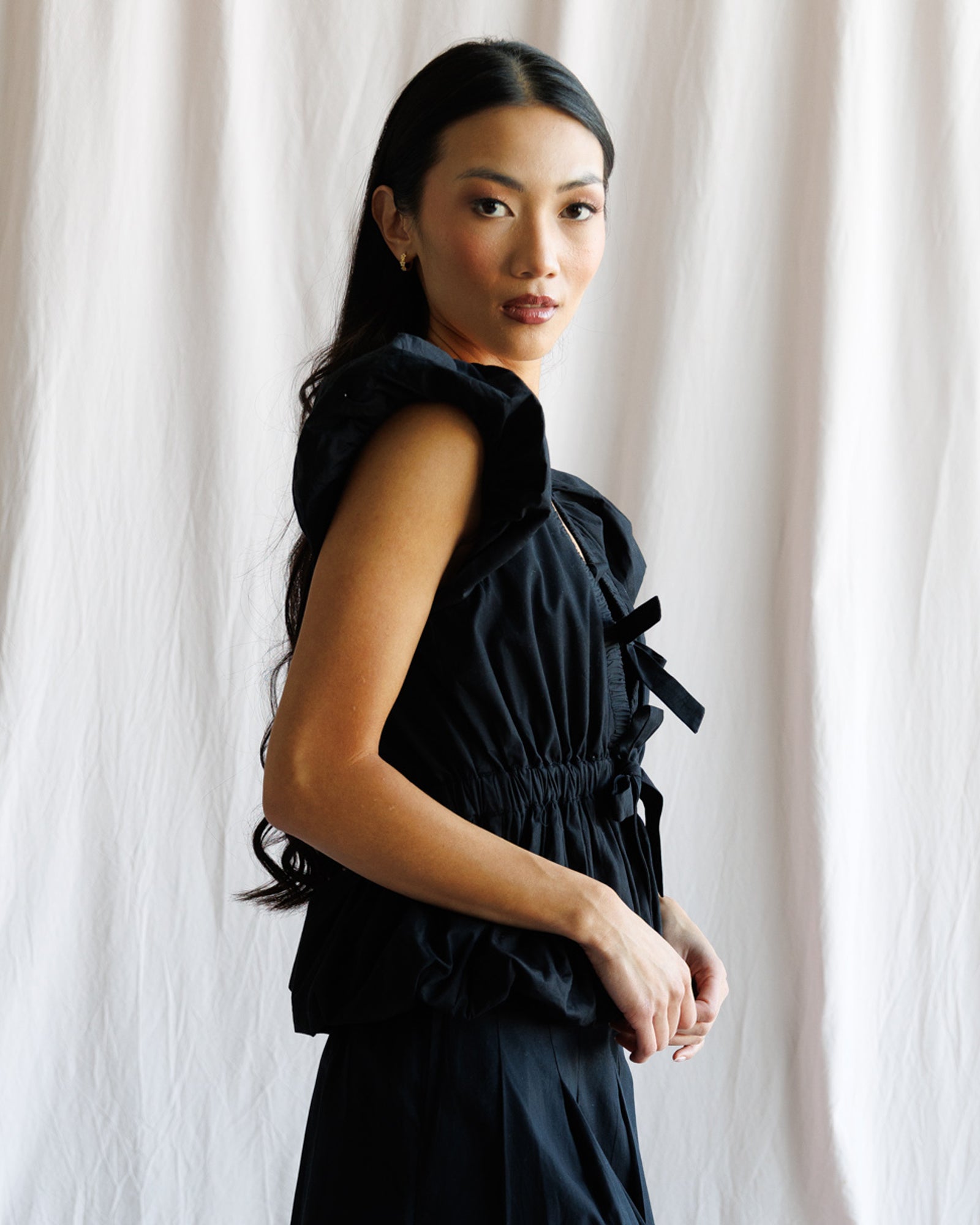 Woman wearing a black sleeveless top with ruched details against a white curtain background