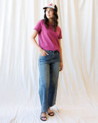 A woman modeling a short-sleeve magenta t-shirt, blue wide-leg jeans, and a baseball cap against a draped white fabric backdrop.
