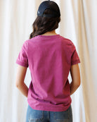A person viewed from behind wearing a short-sleeve crewneck t-shirt in a washed magenta-pink color, paired with blue jeans and a black baseball cap, set against a soft beige curtain background.