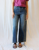 A person wearing medium-wash blue wide-leg denim jeans, paired with a pink top and black sandals, standing against a draped white fabric backdrop.