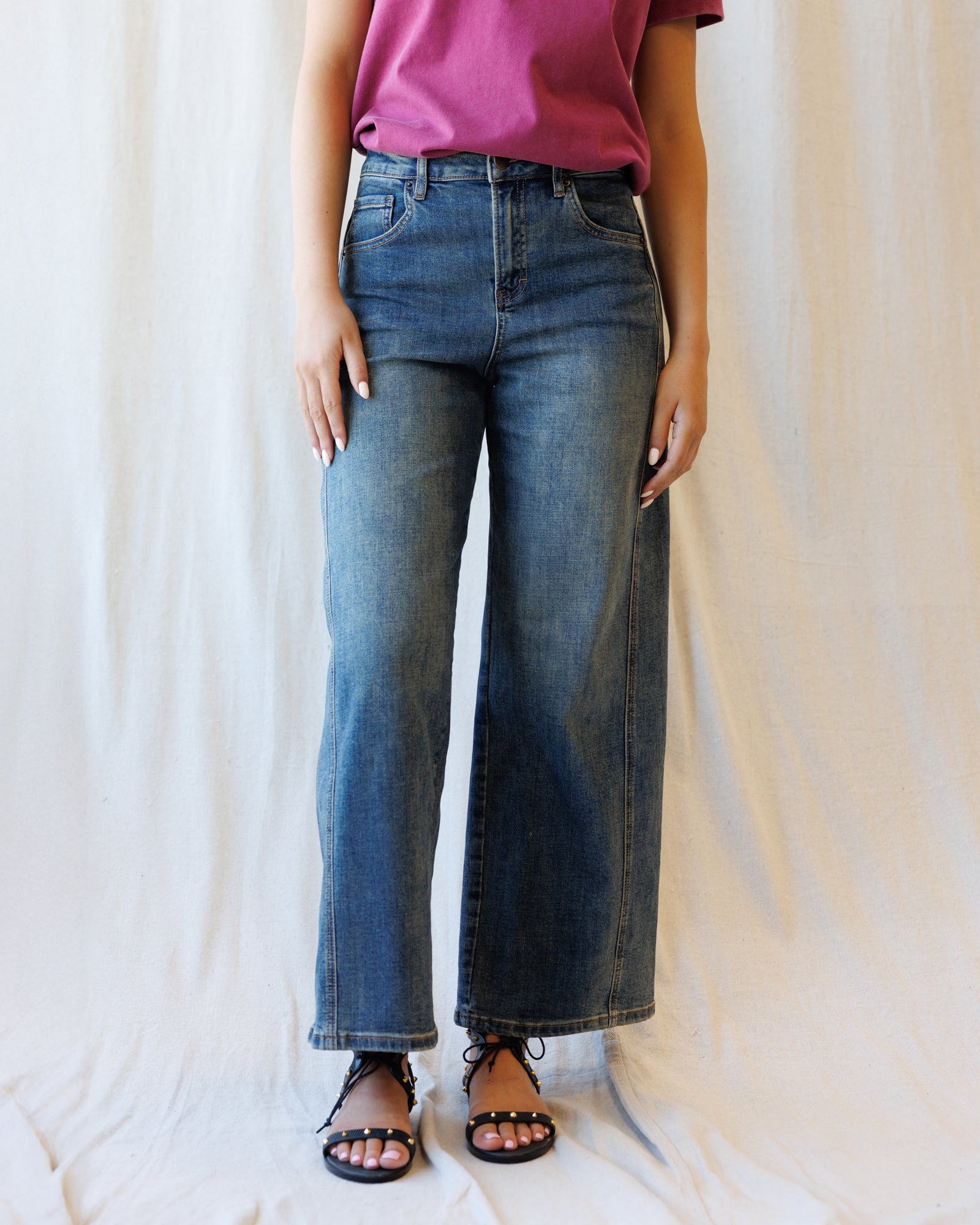 A person wearing medium-wash blue wide-leg denim jeans, paired with a pink top and black sandals, standing against a draped white fabric backdrop.
