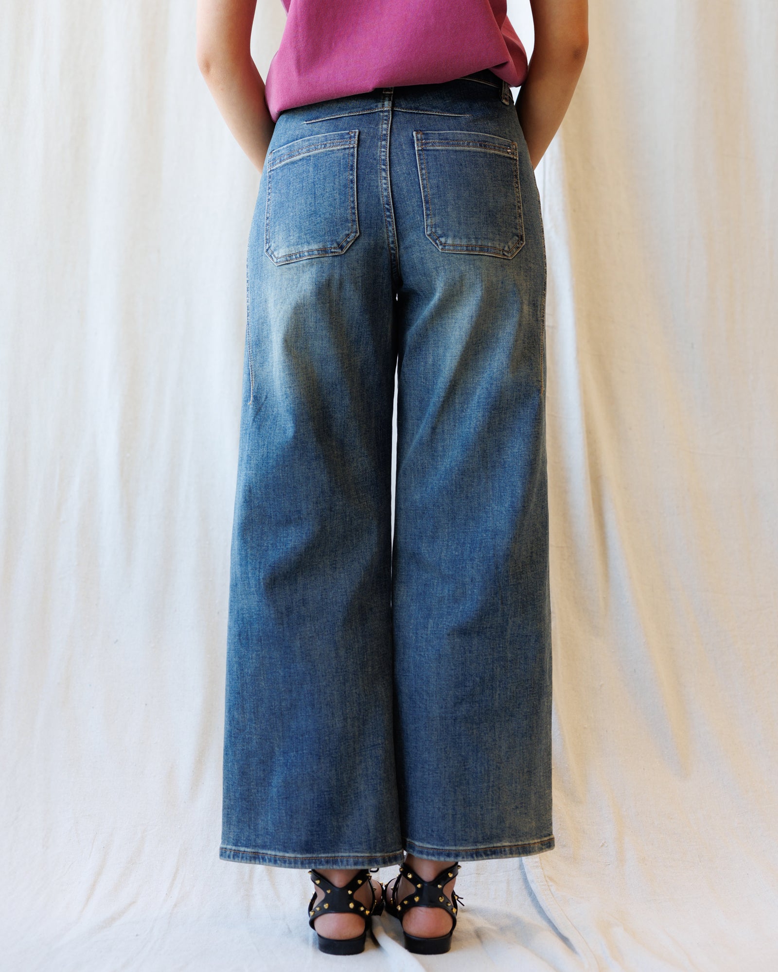 A person shown from the back wearing wide-leg blue denim jeans with a pink top and black strappy sandals, standing against a white fabric backdrop.