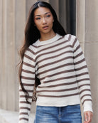 A woman wearing a brown and cream striped knit sweater with blue jeans, standing against a neutral stone wall.