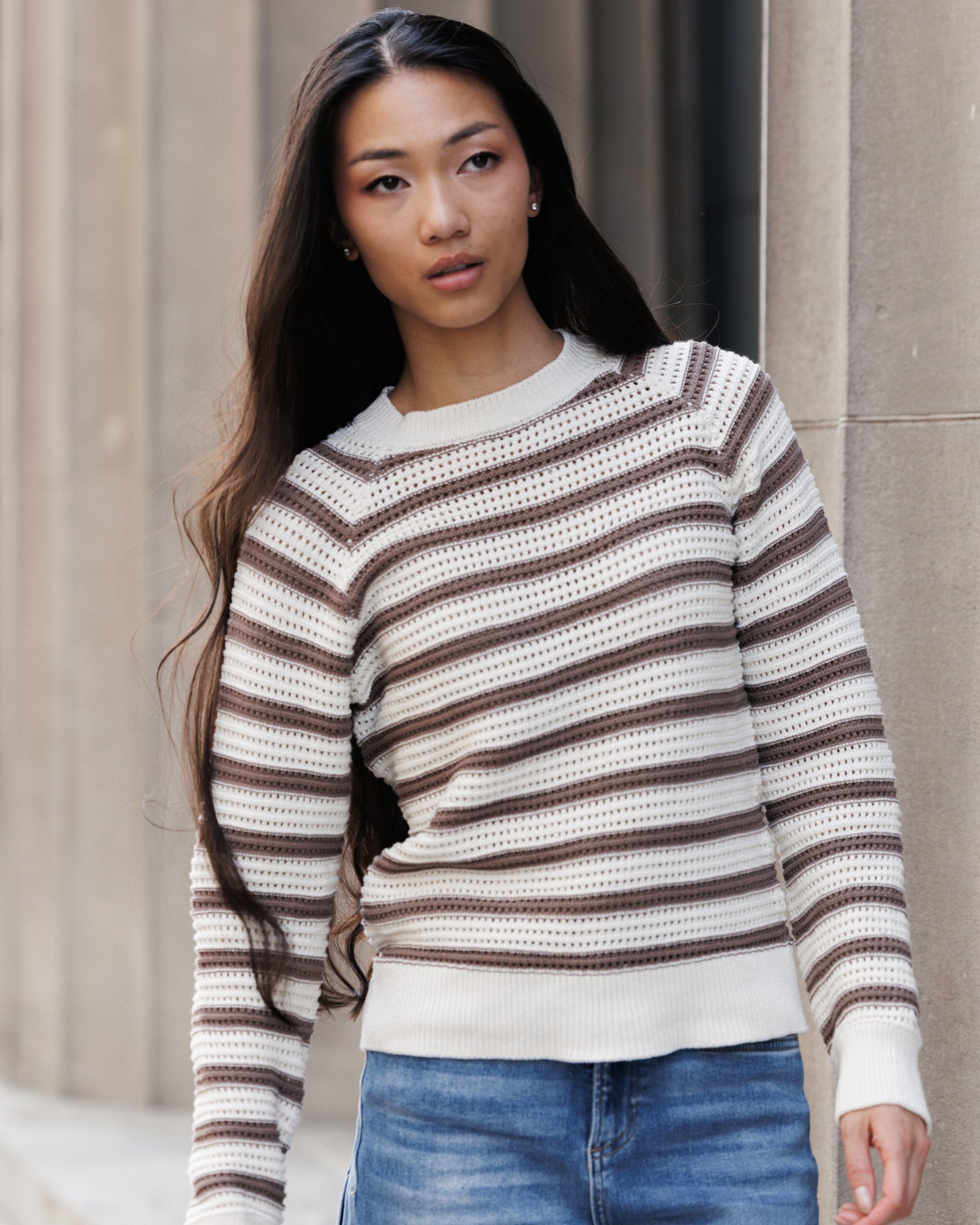 A woman wearing a brown and cream striped knit sweater with blue jeans, standing against a neutral stone wall.