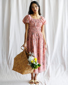 Woman in a pink floral dress holding a woven bag and flowers against a white background