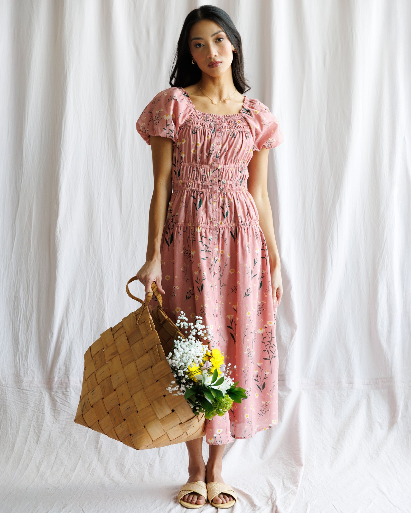 Woman in a pink floral dress holding a woven bag and flowers against a white background