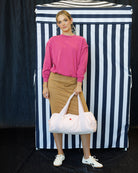A woman stands in front of a black and white vertically striped backdrop, wearing a bright pink long-sleeve top, a tan pencil skirt, and white sneakers, while holding a large pale pink duffel bag with white handles.
