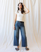 A woman modeling a pair of wide-leg blue jeans with a black belt, paired with a cream t-shirt and floral flat shoes, standing against a draped white fabric backdrop.