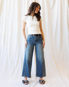 A woman modeling a pair of wide-leg blue denim jeans with a black belt, styled with a white t-shirt and floral print slippers against a draped white fabric backdrop.