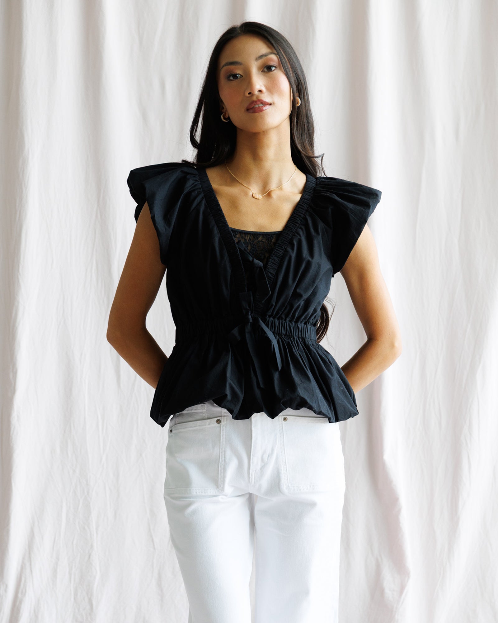 Woman wearing a black top with ruffled shoulders and white pants against a white curtain background