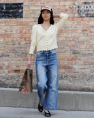 A woman stands against a brick wall wearing a cream button-front cardigan, wide-leg blue jeans, black sandals, and a baseball cap, while holding a shopping bag.