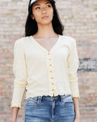 A woman wearing a cream-colored, long-sleeve button-up cardigan top with ruffle trim and frayed edges, paired with blue jeans and a black cap, standing against a brick wall.