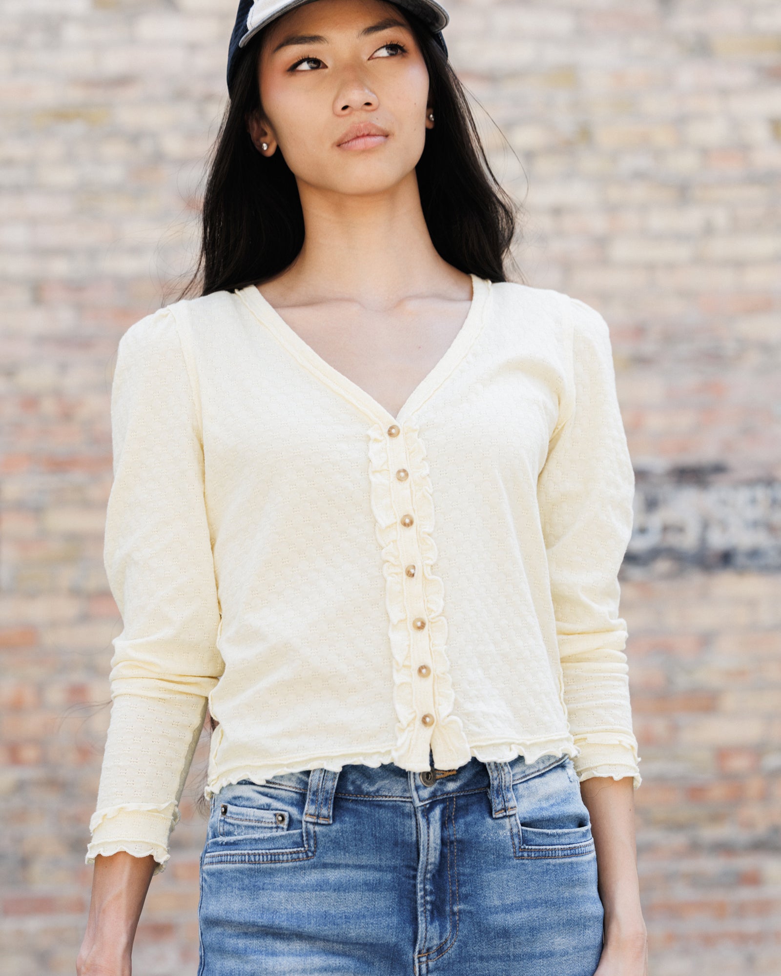 A woman wearing a cream-colored, long-sleeve button-up cardigan top with ruffle trim and frayed edges, paired with blue jeans and a black cap, standing against a brick wall.
