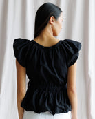 Woman wearing a black blouse with ruffled sleeves against a white curtain background