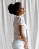 Woman in a light pink sweater and white pants standing against a white curtain.