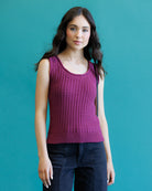 A woman modeling a sleeveless, scoop-neck knit top in a textured magenta-purple color, paired with dark jeans, set against a solid teal background.