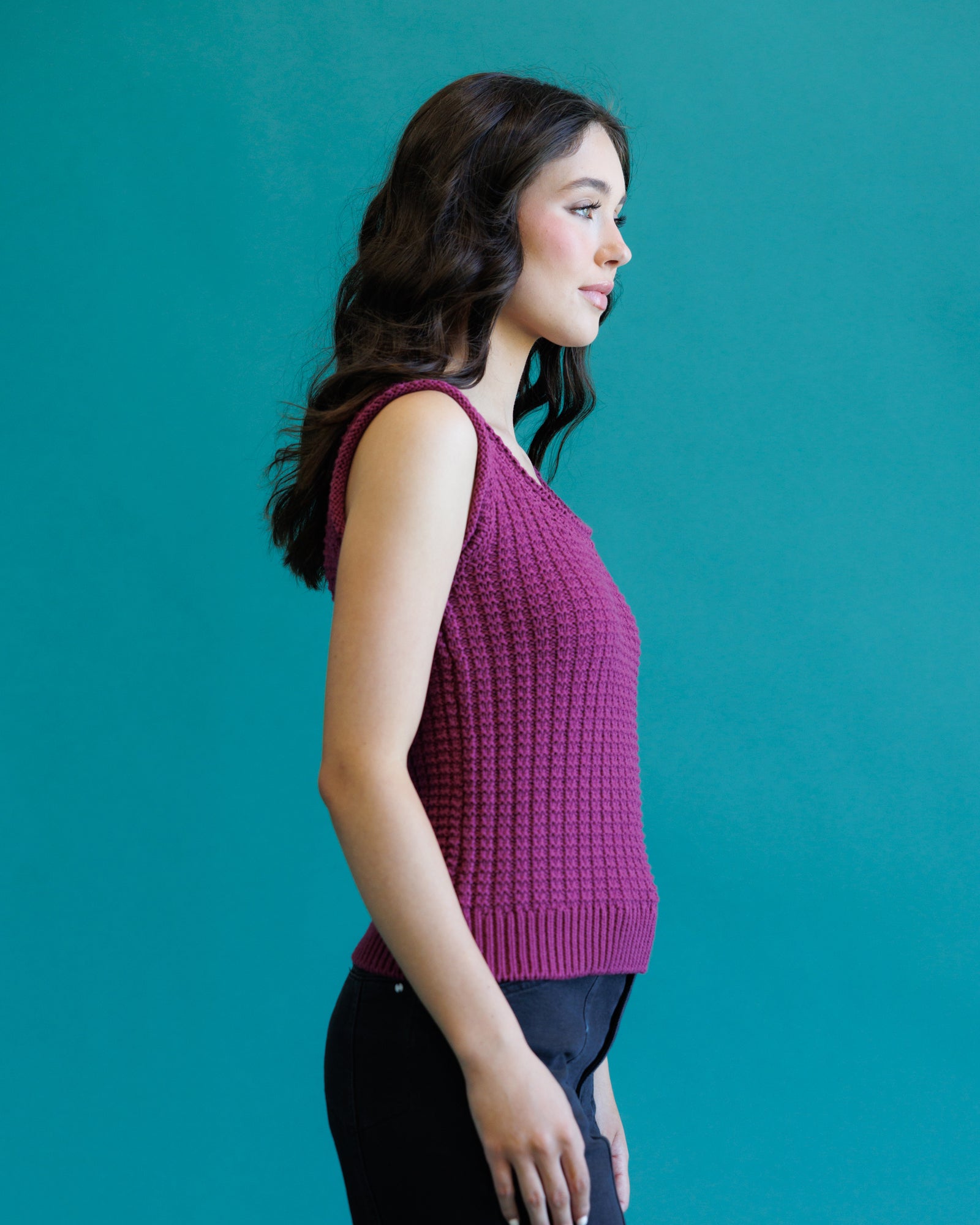 A woman in profile wearing a sleeveless, textured knit top in a deep berry purple, paired with black pants, set against a solid teal background.