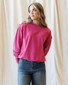 A woman wearing a bright pink long-sleeve crewneck sweatshirt and blue jeans, standing in front of a draped cream fabric backdrop.