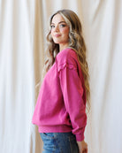 A woman with long wavy light brown hair is shown from the side, wearing a bright pink long-sleeve sweatshirt with ruffle details on the shoulders and blue jeans, standing in front of a draped cream fabric backdrop.