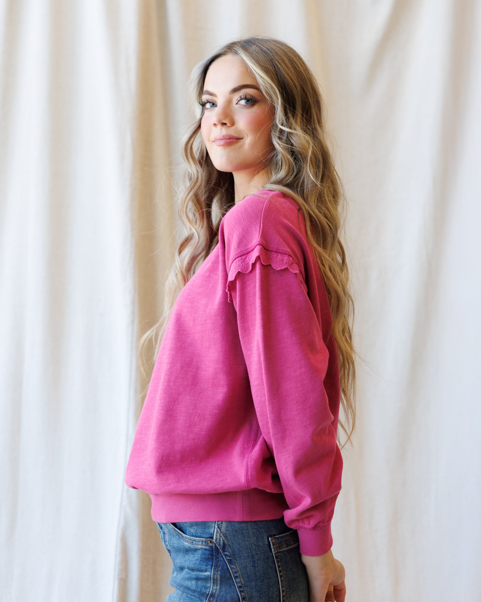 A woman with long wavy light brown hair is shown from the side, wearing a bright pink long-sleeve sweatshirt with ruffle details on the shoulders and blue jeans, standing in front of a draped cream fabric backdrop.