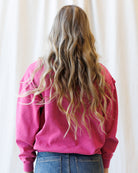 A person viewed from behind wearing a bright pink crewneck sweatshirt and blue jeans, with long, wavy blonde hair, set against a soft white curtain background.