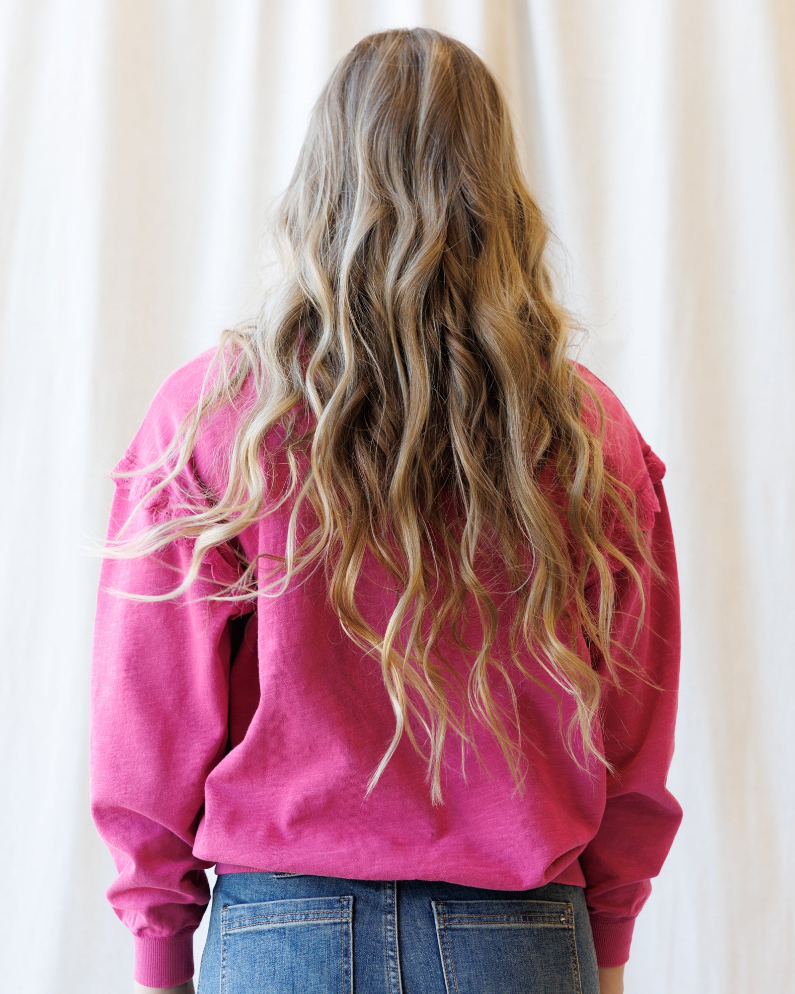 A person viewed from behind wearing a bright pink crewneck sweatshirt and blue jeans, with long, wavy blonde hair, set against a soft white curtain background.
