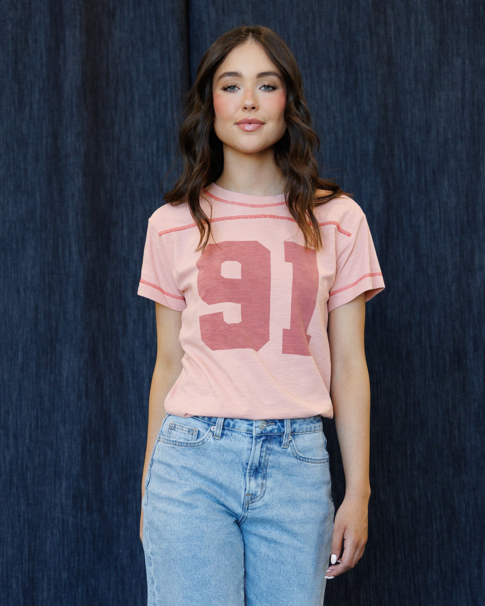 A woman wearing a light pink short-sleeve t-shirt with a large distressed dark pink '91' graphic on the front, paired with light blue jeans, standing against a dark blue textured backdrop.