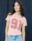 A woman wearing a light pink short-sleeve t-shirt with a large dusty rose '91' graphic on the front, paired with light blue jeans, set against a dark textured backdrop.
