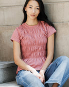 A woman sitting on concrete steps wearing a dusty rose pink smocked short-sleeve t-shirt and blue jeans.