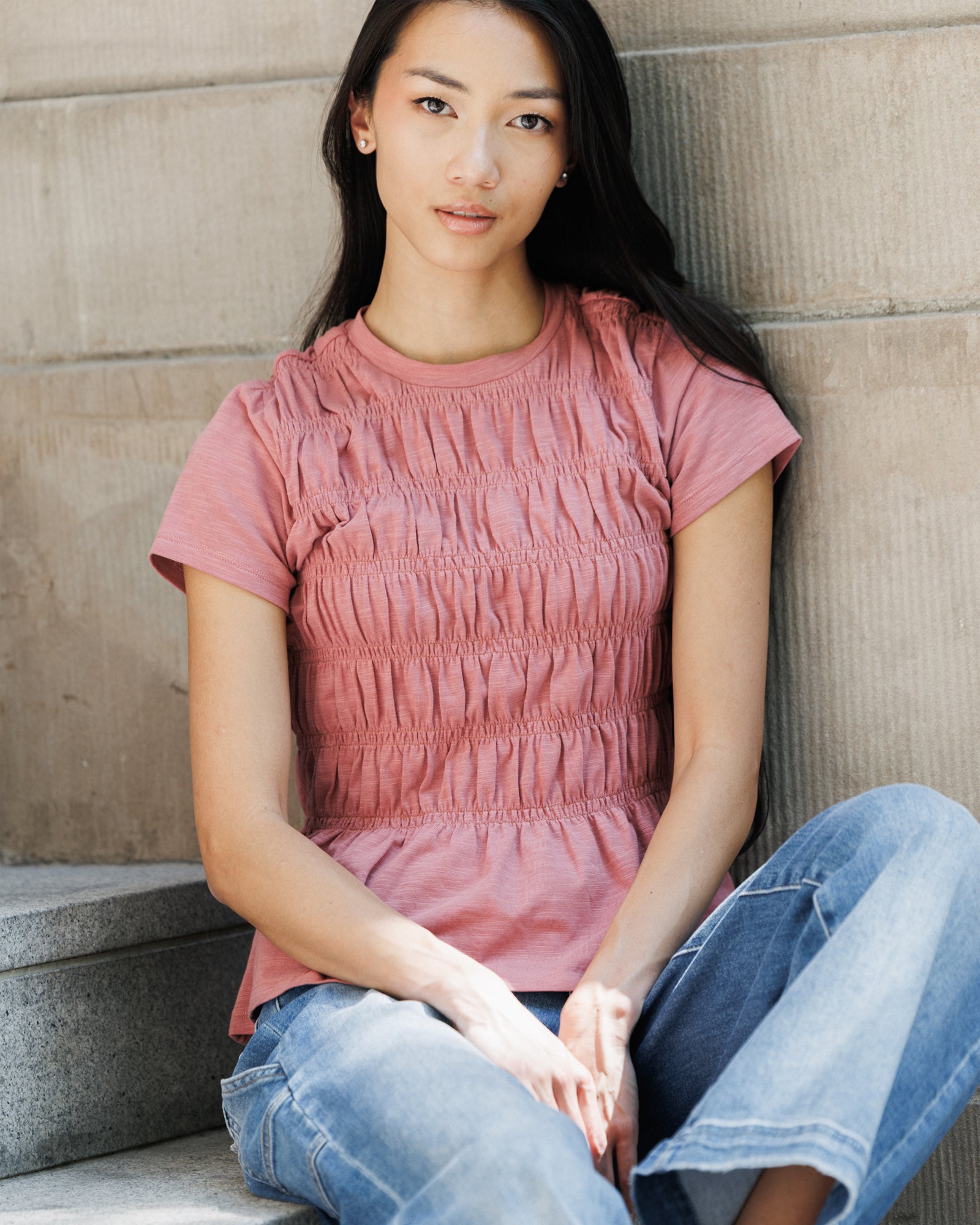 A woman sitting on concrete steps wearing a dusty rose pink smocked short-sleeve t-shirt and blue jeans.