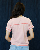 A person viewed from the back wearing a light pink short-sleeve t-shirt with contrasting coral-pink horizontal stripe details across the upper back and sleeve cuffs, paired with light blue jeans against a dark blue textured backdrop.