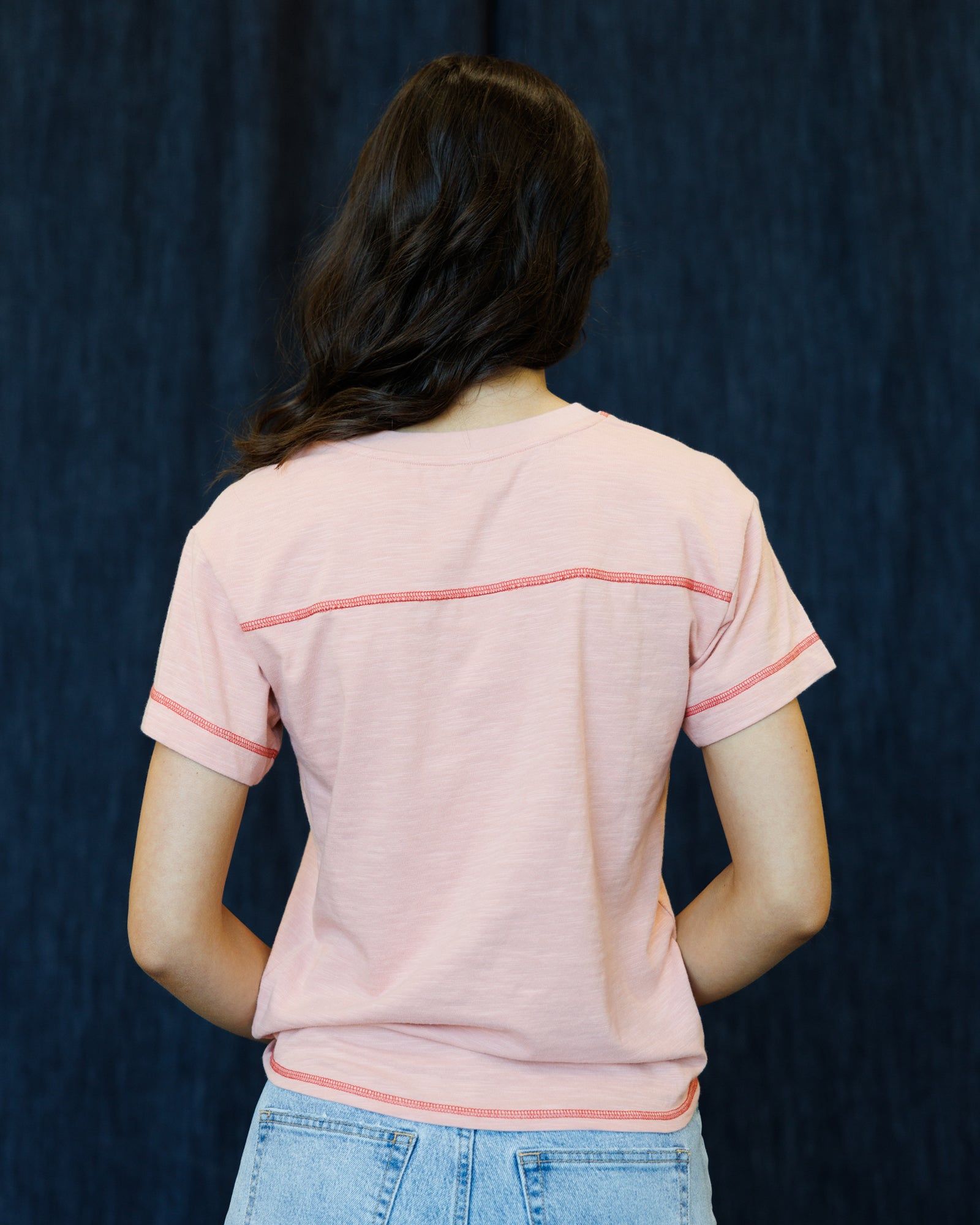 A person viewed from the back wearing a light pink short-sleeve t-shirt with contrasting coral-pink horizontal stripe details across the upper back and sleeve cuffs, paired with light blue jeans against a dark blue textured backdrop.
