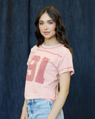 A young woman with wavy brown hair wearing a light pink short-sleeve t-shirt with a distressed dark pink graphic and light blue jeans, standing against a dark blue textured backdrop.