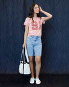 A woman stands against a dark blue textured backdrop wearing a light pink graphic T-shirt with a large pink '91' print, light blue denim shorts, white sneakers, and holding a white tote bag with black handles.