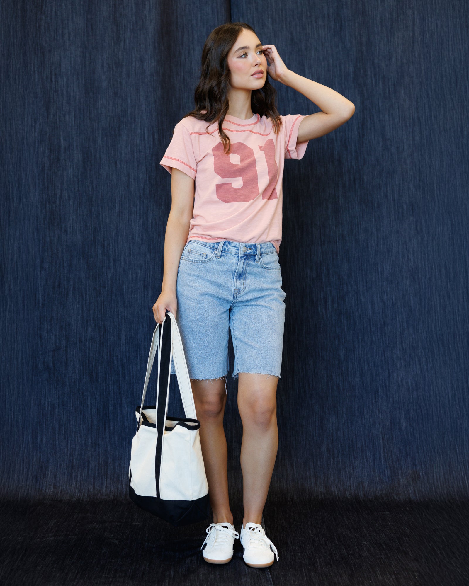 A woman stands against a dark blue textured backdrop wearing a light pink graphic T-shirt with a large pink '91' print, light blue denim shorts, white sneakers, and holding a white tote bag with black handles.