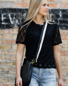 A woman wearing a black short-sleeve lace top and light blue jeans, carrying a black crossbody bag with a white strap, standing against a brick wall.