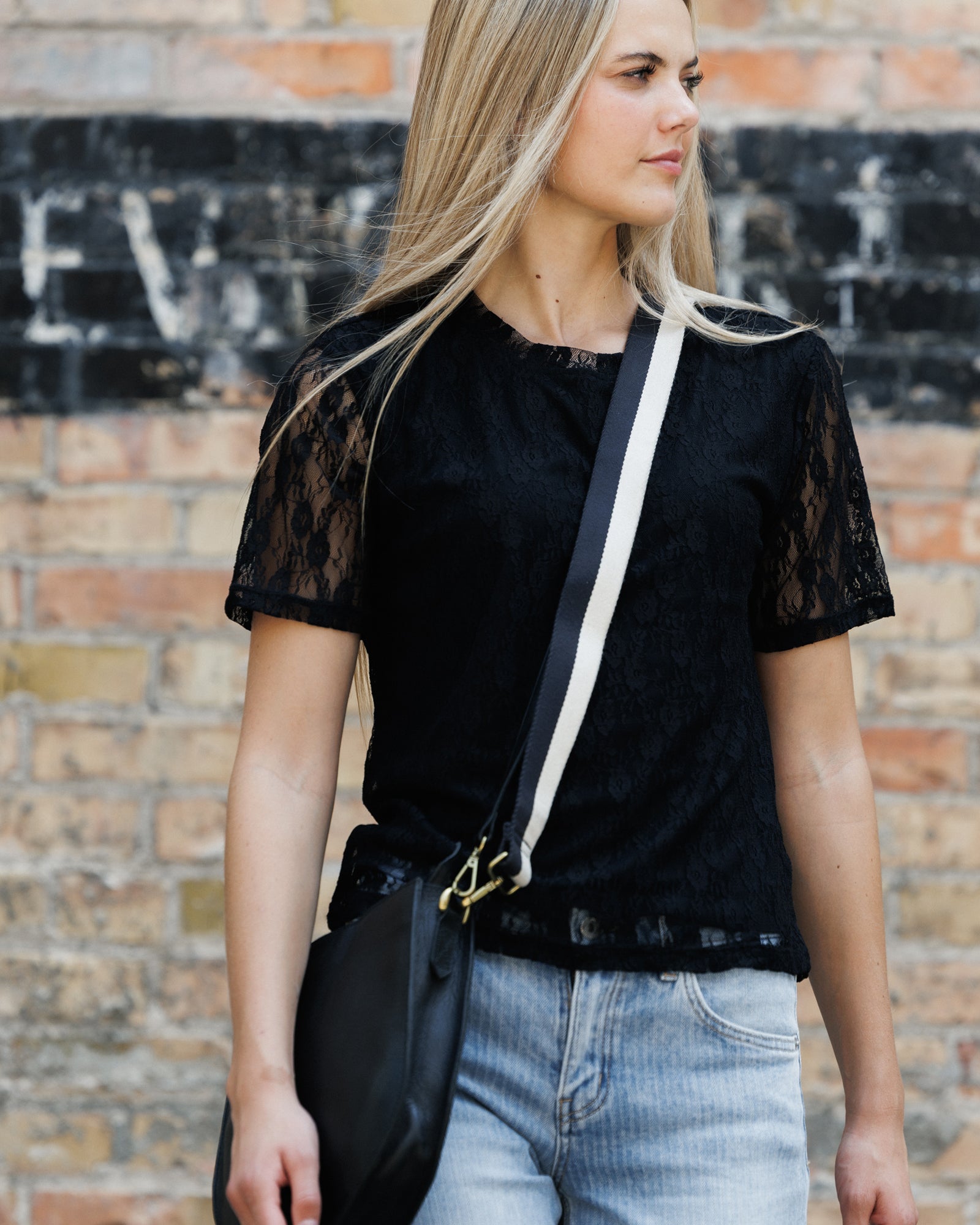 A woman wearing a black short-sleeve lace top and light blue jeans, carrying a black crossbody bag with a white strap, standing against a brick wall.
