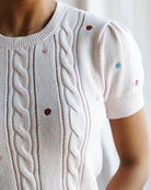 Close-up of a white knit sweater with cable patterns and embroidered flowers.
