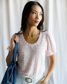 Woman wearing a light pink textured top holding a denim bag against a white curtain background